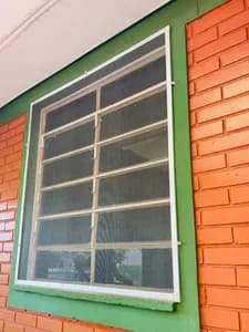 Tela mosquiteiro de alumínio branco instalada com tramelas em janela sobre parede de tijolinho. - UauTelas Terenos MS